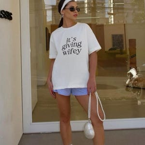 Op de afbeelding: Een wit T-shirt met de tekst "it's giving wifey" erop gedrukt. De persoon draagt een blauw-wit gestreepte short, witte sokken en witte sneakers. Ze draagt ook een witte parelentas.