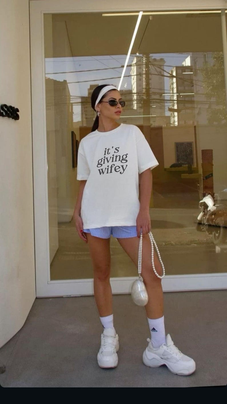 Op de afbeelding: Een wit T-shirt met de tekst "it's giving wifey" erop gedrukt. De persoon draagt een blauw-wit gestreepte short, witte sokken en witte sneakers. Ze draagt ook een witte parelentas.