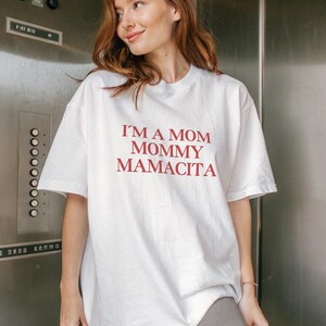 May include: White t-shirt with the text "I'M A MOM MOMMY MAMACITA" in red. The shirt is a casual, oversized style. The model is wearing gray pants.