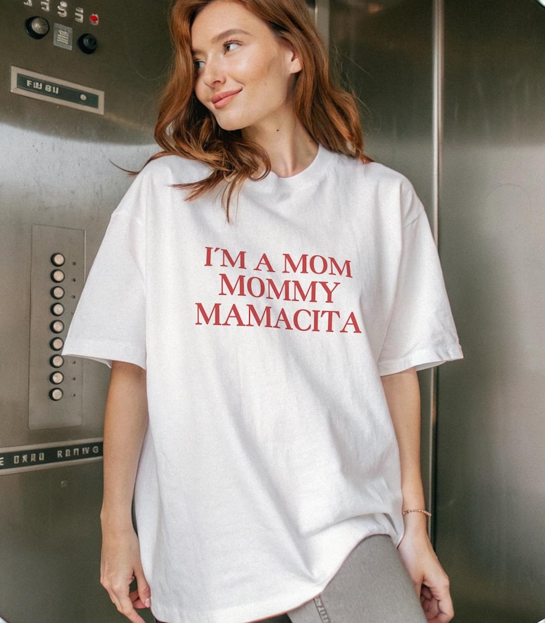 May include: White t-shirt with the text "I'M A MOM MOMMY MAMACITA" in red. The shirt is a casual, oversized style. The model is wearing gray pants.