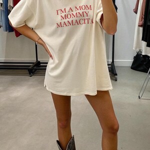 May include: Cream-colored t-shirt with the text "I'M A MOM MOMMY MAMACITA" in red. The shirt is worn with a gold necklace and cowboy boots. The shirt is a casual, oversized fit.