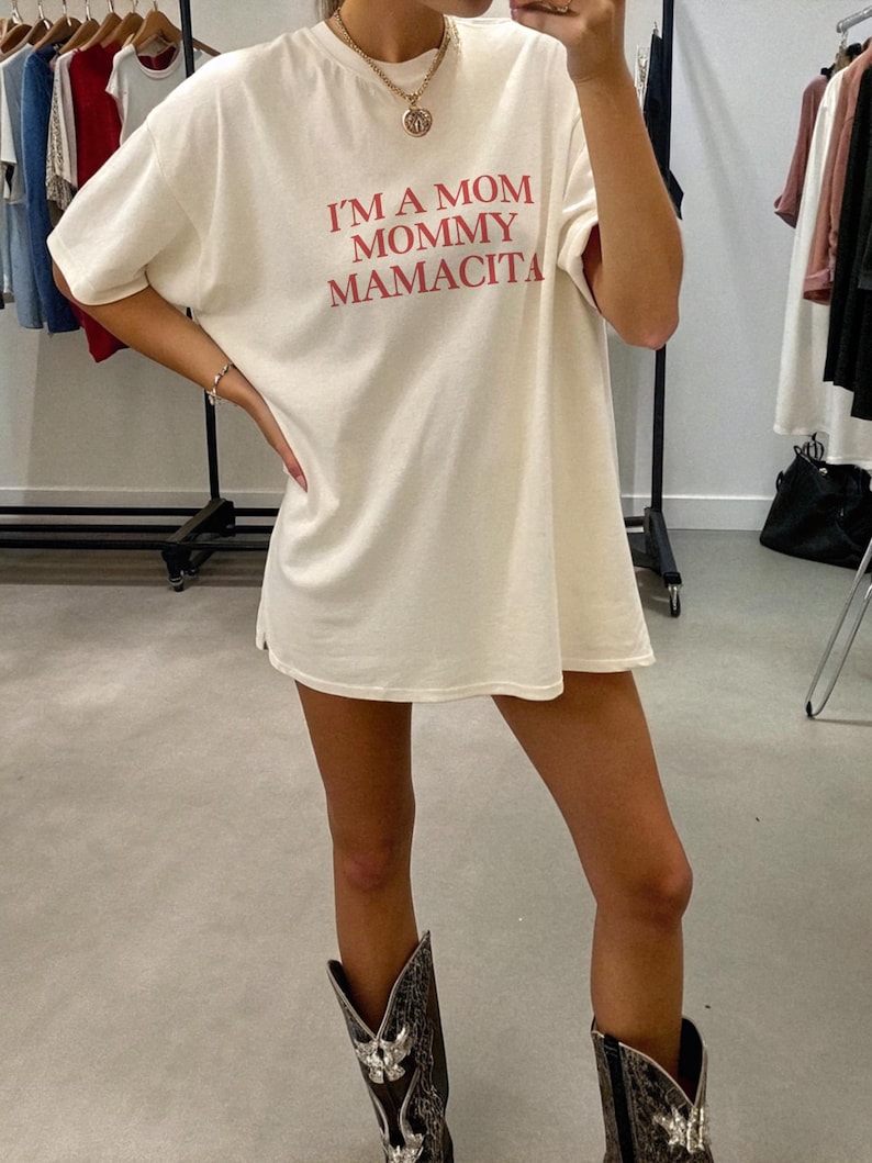 May include: Cream-colored t-shirt with the text "I'M A MOM MOMMY MAMACITA" in red. The shirt is worn with a gold necklace and cowboy boots. The shirt is a casual, oversized fit.