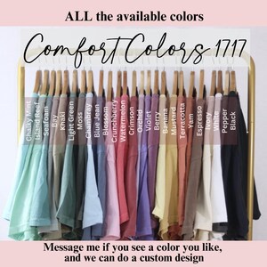 Puede incluir: Exhibici&oacute;n de camisetas Comfort Colors 1717 en varios colores, incluyendo Chalky Mint, Island Reef y Negro, colgadas en una percha dorada. El texto "ALL the available colors" y "Comfort Colors 1717" es visible.