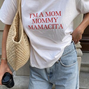 May include: White t-shirt with the red text "I'M A MOM MOMMY MAMACITA." The shirt is paired with light wash blue jeans and a woven beige bag. A black cup is held in the hand.