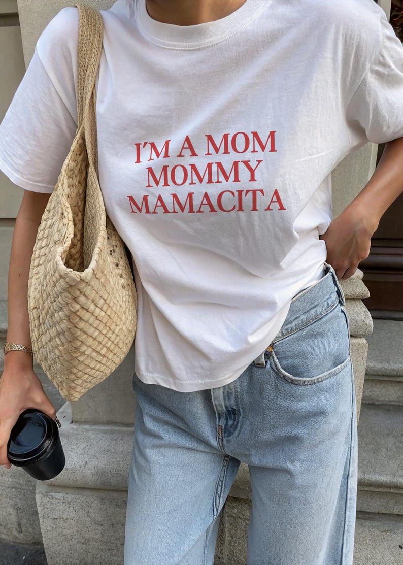 May include: White t-shirt with the red text "I'M A MOM MOMMY MAMACITA." The shirt is paired with light wash blue jeans and a woven beige bag. A black cup is held in the hand.