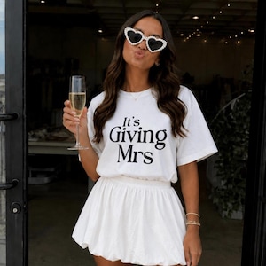 May include: A white short-sleeved t-shirt with the black text It's Giving Mrs printed on the front, paired with a white mini bubble skirt. The outfit is accessorized with heart-shaped sunglasses featuring a white pearl-like trim around the frames.