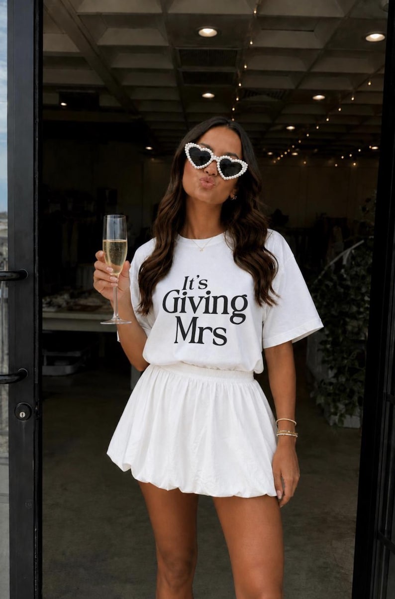 May include: A white short-sleeved t-shirt with the black text It's Giving Mrs printed on the front, paired with a white mini bubble skirt. The outfit is accessorized with heart-shaped sunglasses featuring a white pearl-like trim around the frames.