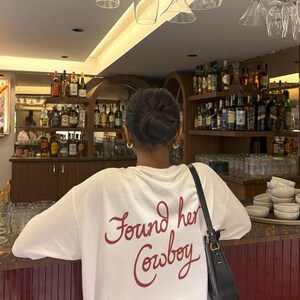 May include: A white t-shirt with the phrase "Found her Cowboy" in red script. The person is standing at a bar with shelves of liquor bottles in the background. A black purse hangs from the person's shoulder.