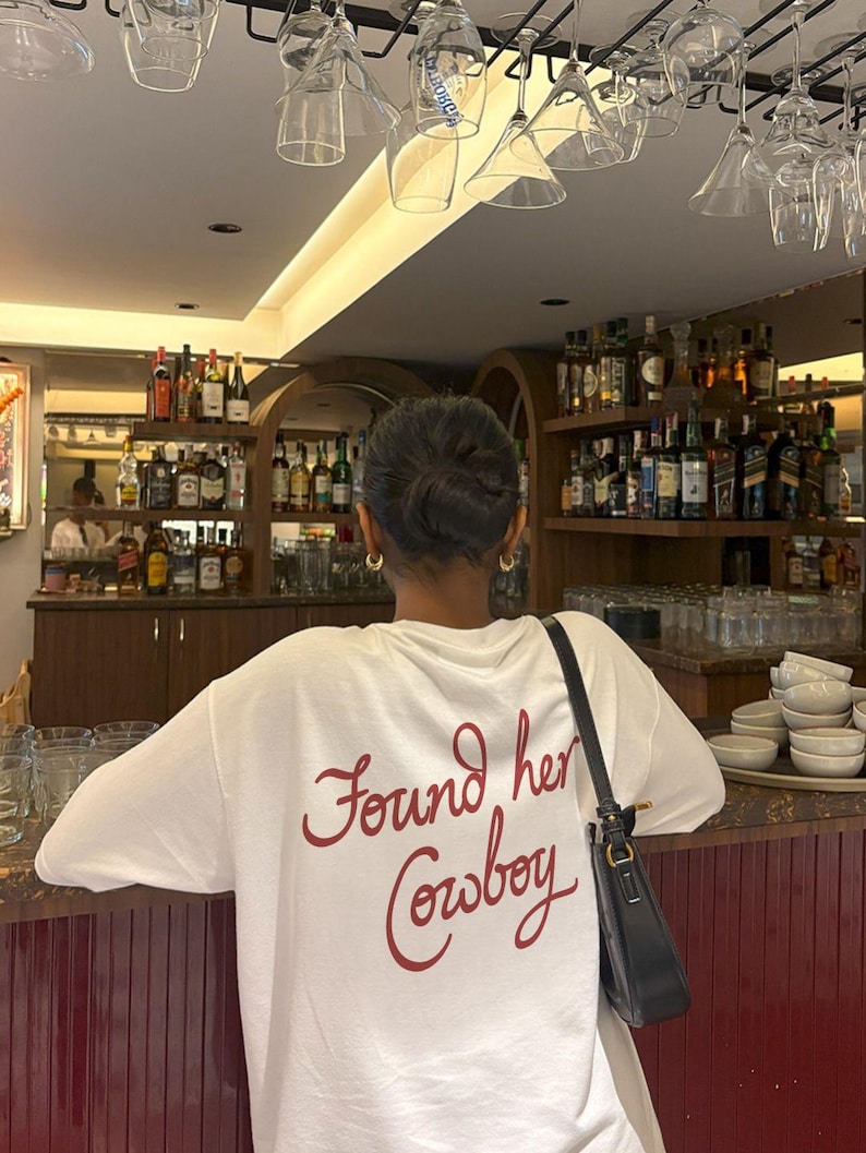 May include: A white t-shirt with the phrase "Found her Cowboy" in red script. The person is standing at a bar with shelves of liquor bottles in the background. A black purse hangs from the person's shoulder.
