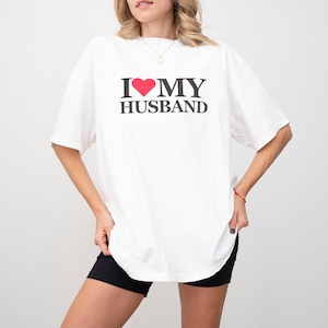 Puede incluir: Camiseta blanca con el texto "I ❤️ MY HUSBAND" en negro y un coraz&oacute;n rojo. La camiseta es de estilo casual, con un corte holgado.