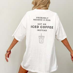 以下が含まれることがあります： 「Probably needed a hug, got an iced coffee instead」というテキストと、アイスコーヒーのカップの絵が描かれた、白のオーバーサイズTシャツです。