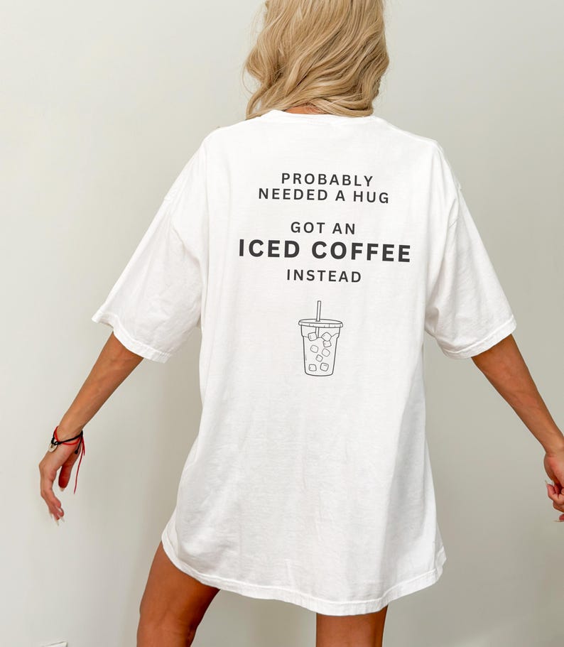 以下が含まれることがあります： 「Probably needed a hug, got an iced coffee instead」というテキストと、アイスコーヒーのカップの絵が描かれた、白のオーバーサイズTシャツです。
