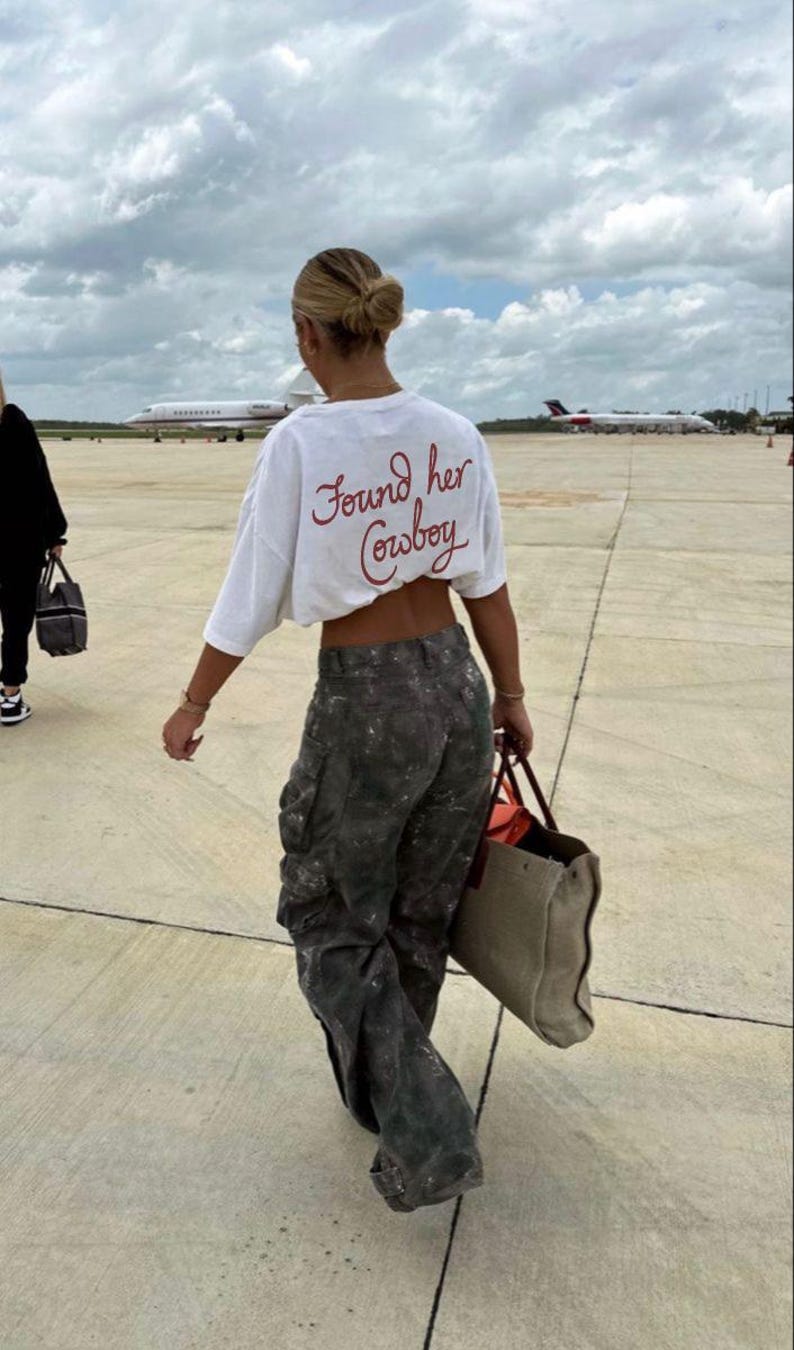May include: A white t-shirt with the text "Found her Cowboy" in red script. The person is wearing gray cargo pants and carrying a beige tote bag. The background includes airplanes and a cloudy sky.