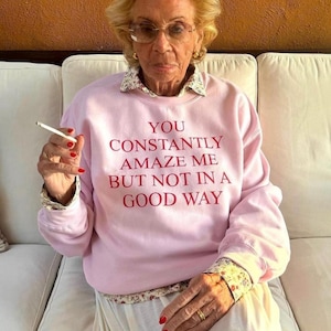 Könnte beinhalten: Hellrosa Sweatshirt mit rotem Text "YOU CONSTANTLY AMAZE ME BUT NOT IN A GOOD WAY". Das Sweatshirt hat einen Kragen und Manschetten mit floralem Muster. Die Person trägt eine Brille und hält eine Zigarette.