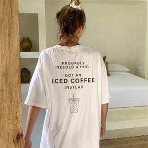 以下が含まれることがあります： 「Probably needed a hug got an iced coffee instead」というテキストと、アイスコーヒーのカップの線画が描かれた、白のオーバーサイズTシャツです。