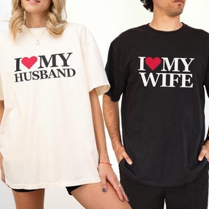 Puede incluir: Dos camisetas a juego, una crema y otra negra. La camiseta crema dice "I ❤️ MY HUSBAND" en texto negro con un coraz&oacute;n rojo. La camiseta negra dice "I ❤️ MY WIFE" con un coraz&oacute;n rojo.