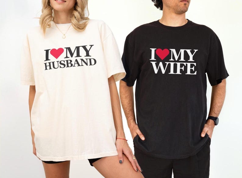 Puede incluir: Dos camisetas a juego, una crema y otra negra. La camiseta crema dice "I ❤️ MY HUSBAND" en texto negro con un coraz&oacute;n rojo. La camiseta negra dice "I ❤️ MY WIFE" con un coraz&oacute;n rojo.