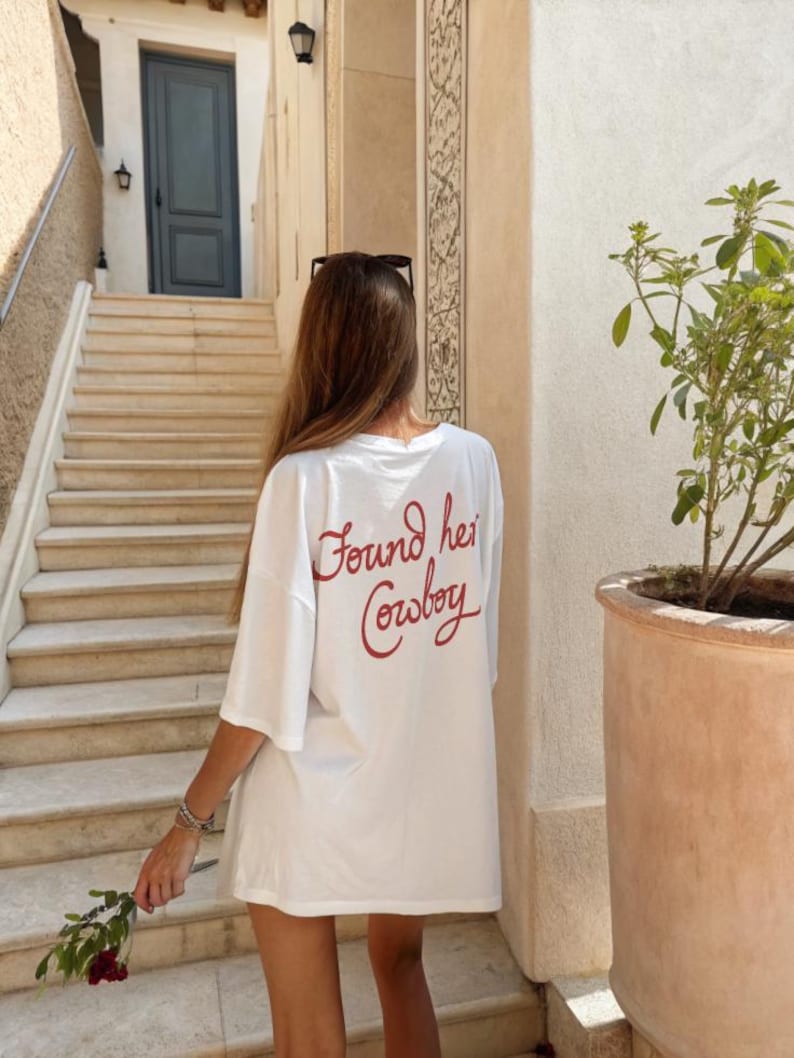 May include: White oversized t-shirt with the red text "Found her Cowboy" on the back. The shirt is worn outdoors, with a person holding a flower. The background includes stairs and a building.