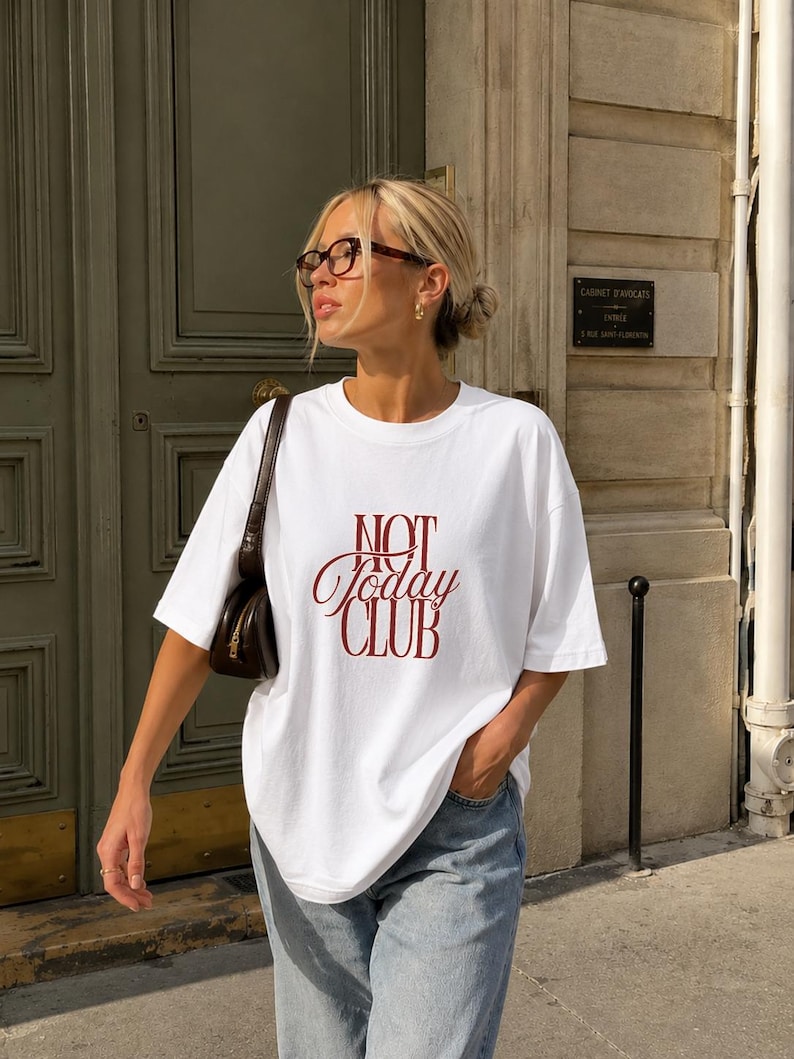 May include: A white short-sleeved t-shirt featuring the dark red text "NOT Today CLUB" printed on the front. The shirt has a crew neckline and a loose, relaxed fit, paired with light blue denim jeans and a dark brown shoulder bag.