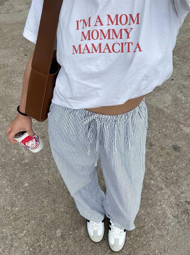 May include: White t-shirt with the text "I'M A MOM MOMMY MAMACITA" in red. The person is wearing blue and white striped pants, white sneakers, and carrying a brown bag and a coffee cup.