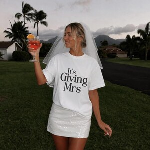 May include: A white short-sleeve t-shirt with the black text It's Giving Mrs printed on the front, paired with a white sequined mini skirt and a sheer white bridal veil. The outfit is styled with a glass containing an orange beverage.