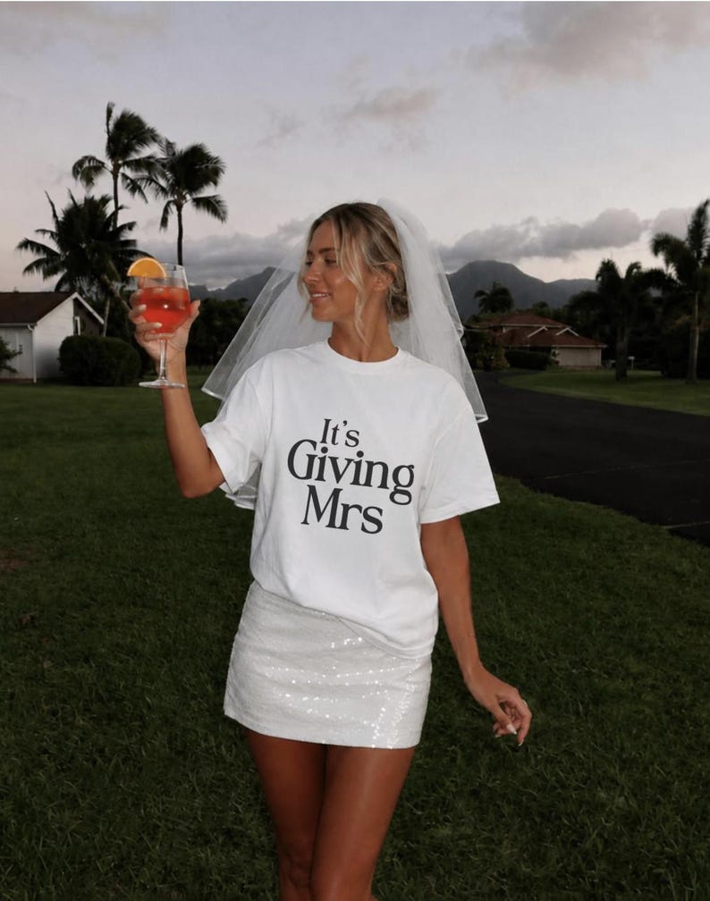 May include: A white short-sleeve t-shirt with the black text It's Giving Mrs printed on the front, paired with a white sequined mini skirt and a sheer white bridal veil. The outfit is styled with a glass containing an orange beverage.