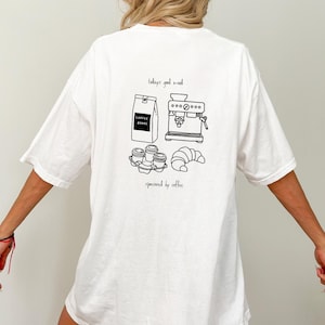 May include: White oversized t-shirt with a black and white illustration of a coffee machine, coffee beans, coffee cups, and a croissant. The text "today's good mood sponsored by coffee" is printed on the shirt.