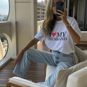 Puede incluir: Camiseta blanca con el texto "I ❤️ MY HUSBAND" en negro y un coraz&oacute;n rojo. La camiseta se combina con vaqueros azul claro. La persona est&aacute; sentada en un sof&aacute; blanco con detalles de madera.