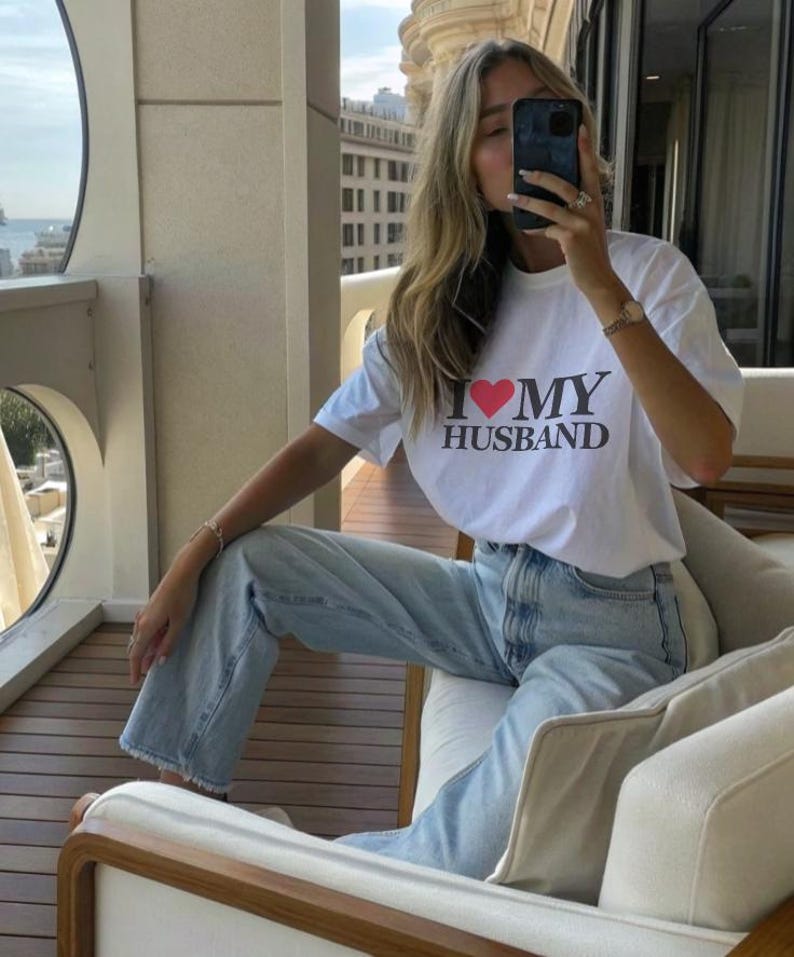 Puede incluir: Camiseta blanca con el texto "I ❤️ MY HUSBAND" en negro y un coraz&oacute;n rojo. La camiseta se combina con vaqueros azul claro. La persona est&aacute; sentada en un sof&aacute; blanco con detalles de madera.