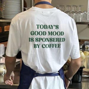 May include: White t-shirt with the text "TODAY'S GOOD MOOD IS SPONSORED BY COFFEE" in green lettering. The shirt is worn by a person wearing a blue apron. The image is taken in a kitchen setting.