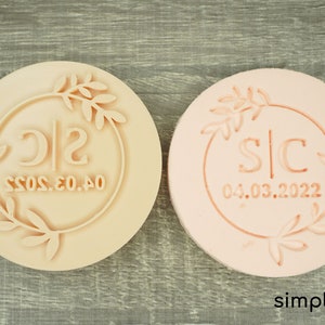 May include: Two round, light-colored stamps with a floral wreath design. One stamp has the initials "SC" and the date "04.03.2022", while the other has "SC" and "04.03.2022". The stamps are on a wooden surface.