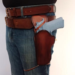 Pode incluir: Um coldre de couro marrom com uma pistola azul claro presa a um cinto de couro marrom. O cinto é usado sobre calças jeans azuis.
