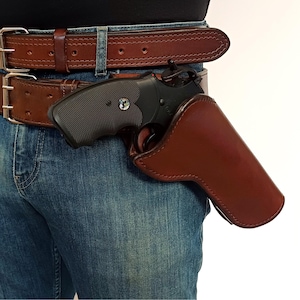 Op de afbeelding: Een bruine leren holster met een zwart pistool erin. De holster wordt gedragen op een bruine leren riem met een zilveren gesp. De persoon draagt een blauwe jeans.