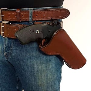 Könnte beinhalten: Ein schwarzer Revolver in einem braunen Lederholster, das an einem dicken braunen Ledergürtel mit einer zweizinkigen Metallschnalle befestigt ist. Der Gürtel wird über einer blauen Jeans getragen und zeigt die glatte Oberfläche des Holsters.