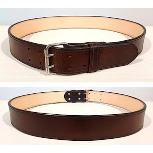 May include: A dark brown leather belt featuring a double-prong silver-toned metal buckle and a light beige interior lining. The belt has two rows of adjustment holes along the strap and a matching leather keeper loop near the buckle assembly.
