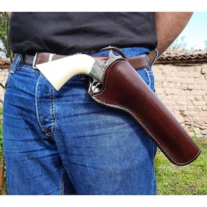Könnte beinhalten: Ein dunkelbraunes Lederholster mit einem silberfarbenen Revolver mit cremefarbenem Griff. Das Holster ist an einem braunen Ledergürtel befestigt, der über einer blauen Jeans getragen wird. Der Revolver zeigt sichtbaren Text am Metallrahmen.