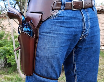 May include: A dark brown leather drop-leg holster secured to a matching brown belt, worn over blue denim jeans. The holster contains a metallic blue revolver with a dark grip, featuring a strap that wraps around the upper thigh for stability.