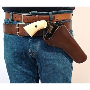May include: A dark brown leather holster containing a revolver with a cream-coloured grip, secured to a wide brown leather belt worn over blue denim jeans. The revolver features a black metal frame and a visible screw on the handle grip.