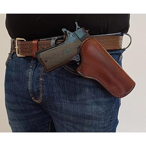 Könnte beinhalten: Ein dunkelbraunes Lederholster mit einer schwarzen halbautomatischen Pistole, befestigt an einem braunen geprägten Ledergürtel über einer blauen Jeans. Das Holster hat sichtbare Nähte an den Kanten und sitzt an der Hüfte.