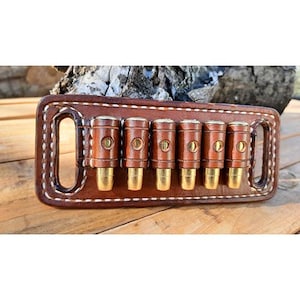 May include: A brown leather cartridge holder featuring white stitching and six individual loops. Each loop contains a brass-cased bullet with a gold-coloured tip. The rectangular leather base includes two side slots for attachment to a belt.