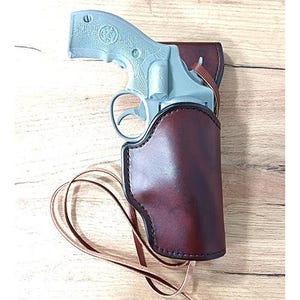 May include: A light grey toy revolver with a textured grip is partially inserted into a dark brown leather holster. The holster features a smooth, polished finish with visible stitching along the edges and includes a thin leather cord attached.