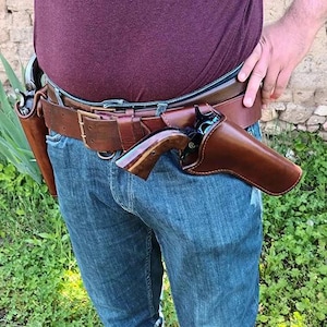 May include: A brown leather gun belt featuring two matching leather holsters, each containing a revolver with a dark metal finish and a polished wooden grip. The belt is secured with a brass-coloured metal buckle and worn over blue denim jeans.