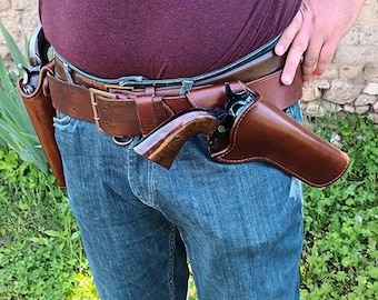 Op de afbeelding: Een bruine lederen wapengordel met twee bijpassende lederen holsters, elk met een revolver met een donkere metalen afwerking en een gepolijste houten greep. De riem is vastgemaakt met een messingkleurige metalen gesp over een blauwe spijkerbroek.