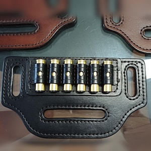 Peut inclure: Un porte-cartouches en cuir noir avec six cartouches à bout en laiton. Le support a une forme rectangulaire avec des bords arrondis et deux découpes ovales. L'arrière-plan montre des parties d'autres articles en cuir.