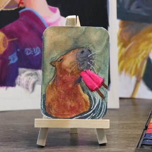 Könnte beinhalten: Ein Aquarellgemälde eines Capybaras, das ein rosa Eis am Stiel genießt, auf einer kleinen Holzstaffelei präsentiert. Das Kunstwerk zeigt das Capybara im Wasser, mit detailliertem Fell und Schnurrhaaren. Der Hintergrund umfasst weitere Gemälde und eine Aquarellpalette.