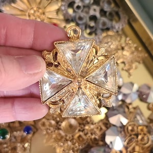 May include: A gold-toned pendant featuring a cross design with four large, clear, triangular-cut crystals. The pendant has ornate gold filigree detailing around the crystals and a loop at the top for hanging. The background shows other vintage jewelry.