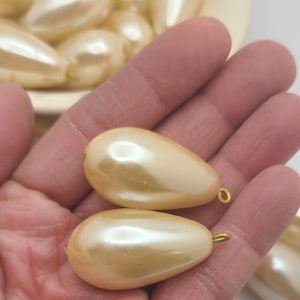 May include: Two large, pearlescent teardrop-shaped beads with gold-colored loops are held in a hand. Several similar beads are visible in the background, suggesting a craft supply or jewelry component. The beads have a smooth, glossy finish.