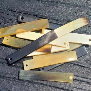 May include: A collection of rectangular, flat beads in various colors, including cream, black, and shades of yellow and brown. Each bead has a small hole at one end, suggesting they are for stringing or crafting.