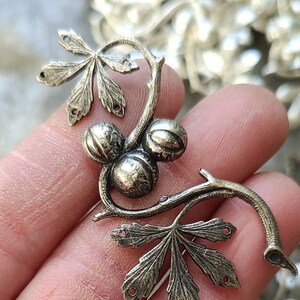 May include: A silver-toned metal charm featuring a stylised branch with three round fruits and two leaves. The charm has holes for attaching it to a piece of jewellery.