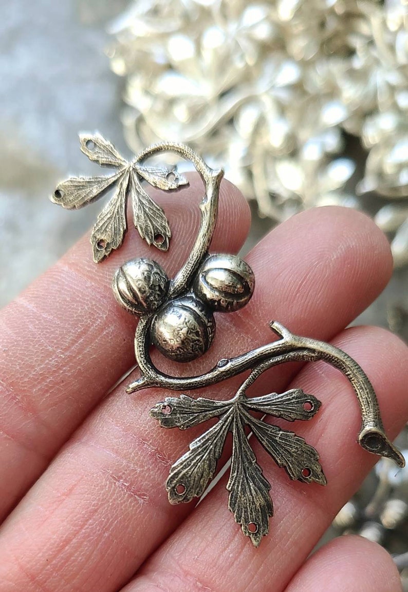 May include: A silver-toned metal charm featuring a stylised branch with three round fruits and two leaves. The charm has holes for attaching it to a piece of jewellery.